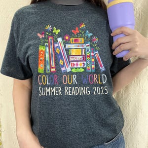May include: A dark grey t-shirt with a colorful illustration of stacked books with flowers and butterflies. The text "Color Our World Summer Reading 2025" is printed below the illustration.