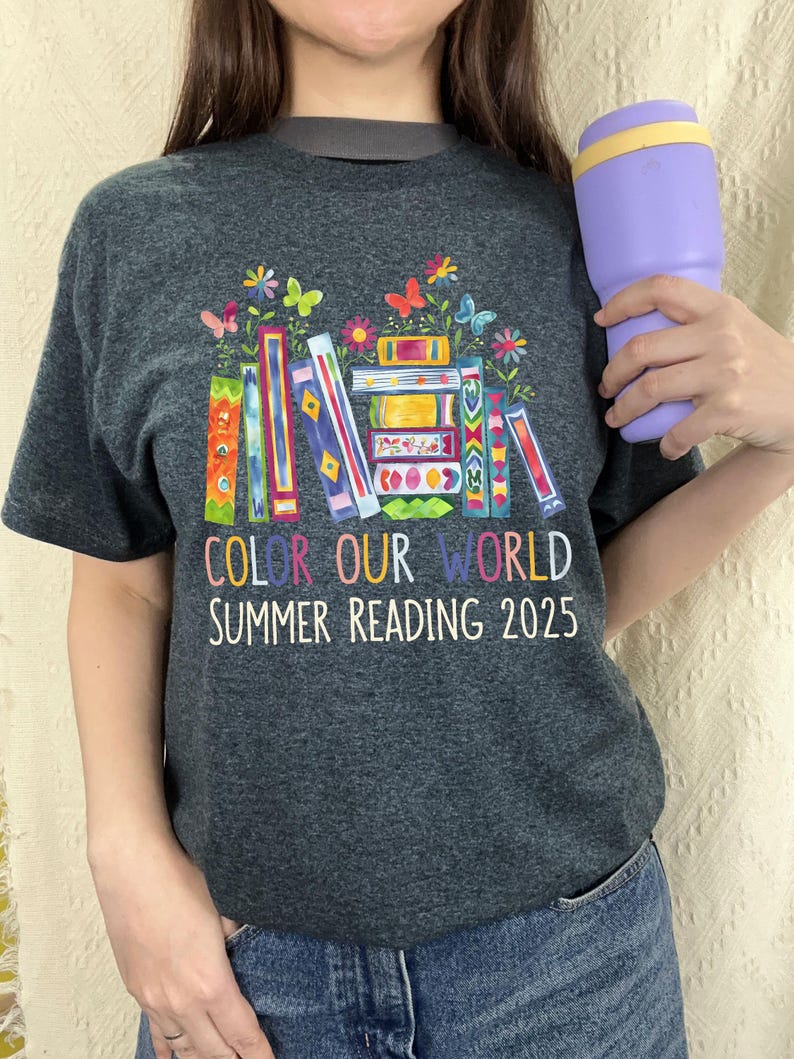 May include: A dark grey t-shirt with a colorful illustration of stacked books with flowers and butterflies. The text "Color Our World Summer Reading 2025" is printed below the illustration.