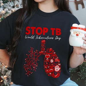 Può includere: Maglietta nera con la scritta rossa "STOP TB" e la scritta bianca "World Tuberculosis Day". La maglietta presenta un disegno polmonare rosso composto da cuori e simboli di mani. Una tazza di Babbo Natale è tenuta in mano.