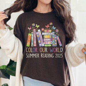 May include: A brown t-shirt with a colorful stack of books and butterflies. The text "Color Our World Summer Reading 2025" is printed on the shirt.