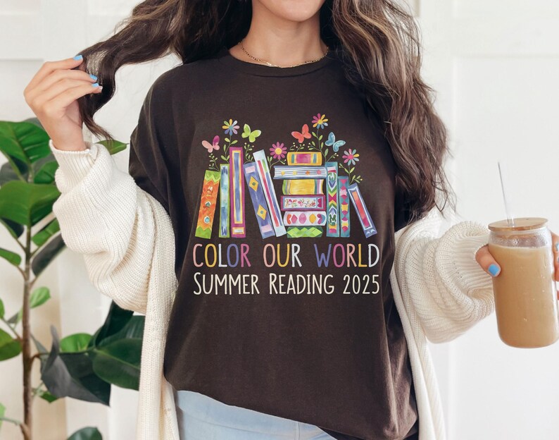 May include: A brown t-shirt with a colorful stack of books and butterflies. The text "Color Our World Summer Reading 2025" is printed on the shirt.