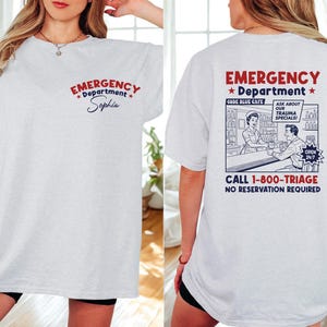 May include: A light gray t-shirt with the words "EMERGENCY Department" in red and blue, and the name "Sophia" in cursive. The back of the shirt features a vintage-style illustration and the text "CALL 1-800-TRIAGE".