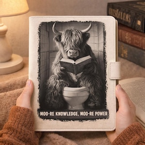 May include: A cream-colored tablet case featuring a black and white image of a Highland cow reading a book while seated on a toilet. The text "MOO-RE KNOWLEDGE, MOO-RE POWER" is printed below the image.