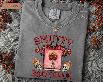 May include: A gray Comfort Colors t-shirt with the words "Smutty Book Club" in red, featuring a book with a flaming rose, roses, and skull graphics. A silver necklace with a pendant rests on the shirt.