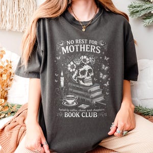 Può includere: T-shirt grigio scuro con la scritta "NO REST FOR MOTHERS BOOK CLUB" e un teschio con una corona floreale. Il design include libri, farfalle, una candela e una tazza da tè. Anche la frase "fueled by coffee, chaos and chapters" è stampata.