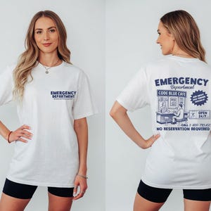 May include: White t-shirt with "Emergency Department" printed on the front and back. The back of the shirt features a graphic for "Code Blue Cafe" with additional text and illustrations. The shirt is worn with black shorts.