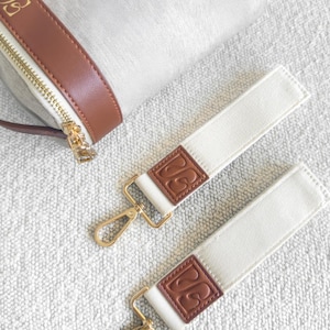 以下が含まれることがあります： ベージュのハンドバッグのクローズアップ。茶色のレザーストラップとゴールドのジッパーが付いています。茶色の革パッチとゴールドの留め金が付いた2本の白いキャンバスストラップも表示されています。革パッチにはエンボス加工のデザインが施されています。