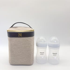 以下が含まれることがあります： ベージュの断熱ベビーボトルバッグ。黒い上部と金色のジッパー、ハンドル、ロゴが付いています。白い乳首が付いた2つの透明なPhilips Aventベビーボトルがバッグの隣にあります。