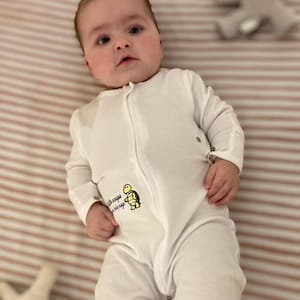 May include: A baby wearing a white, long-sleeved sleepsuit with a full-length zipper. The sleepsuit features a small graphic of a turtle and the words "little snaps, big nap". The baby is lying on a striped sheet.