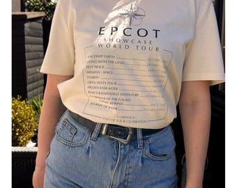 May include: Cream-colored EPCOT Showcase World Tour t-shirt with a list of attractions and dates. The shirt is paired with denim shorts and a black belt. The text "SHOWCASE WORLD TOUR" is at the top. The Parks Tour Collection, July 5th 2024.