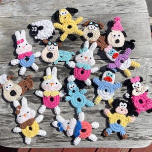 以下が含まれることがあります： カラフルな手作りのキャラクターかぎ針編みアップリケのコレクション。アップリケは、ウサギ、犬、アヒルなど、さまざまな動物や漫画のキャラクターのデザインを特徴としています。各キャラクターは、帽子やリボンなどの詳細を備えた、独自の配色とデザインを持っています。