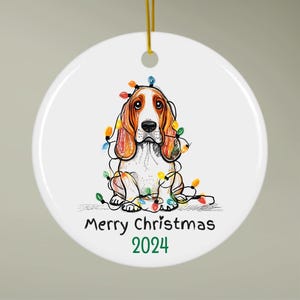 Op de afbeelding: Witte, ronde kerstbal met een afbeelding van een Basset Hound, versierd met kleurrijke kerstverlichting. De tekst "Merry Christmas 2024" staat eronder. Een gouden touwtje is bevestigd om op te hangen.