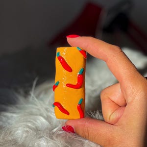 Peut inclure: Un petit objet rectangulaire jaune décoré de piments rouges avec des tiges bleues. L'objet est tenu par une main avec du vernis à ongles rouge. L'arrière-plan est flou, avec une texture douce, blanche et duveteuse.