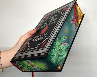 May include: A hardcover book titled "Fire and Blood" by George R.R. Martin. The black cover features white and red accents, with a decorative border. The open book reveals a green dragon illustration and a red dragon. A red ribbon bookmark is visible.