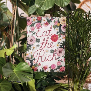 May include: A white garden flag with a floral pattern and the text "Eat the Rich" in red cursive lettering.