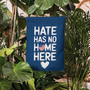 May include: A blue garden flag with white text that reads "Hate Has No Home Here". The text is in a brushstroke font. There is a small American flag heart in the middle of the text. There is a white heart at the bottom of the flag.