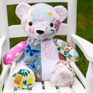 May include: A patchwork teddy bear constructed from various fabric patterns including blue butterflies, pink silhouettes, citrus fruits, and floral prints. The bear features black button eyes, a black nose, and a pink embroidered monogram on the chest.