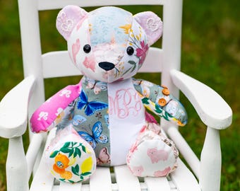 Könnte beinhalten: Ein Patchwork-Teddybär aus verschiedenen Stoffmustern, darunter blaue Schmetterlinge, rosa Silhouetten, Zitrusfrüchte und Blumenmuster. Der Bär hat schwarze Knopfaugen, eine schwarze Nase und ein aufgesticktes rosa Monogramm auf der Brust.