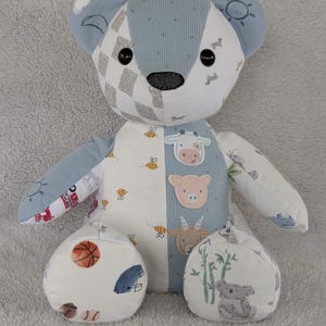 May include: A patchwork teddy bear with a variety of patterned fabrics in blue, white, and gray. The bear features a gray nose, black button eyes, and appliqued animal faces. The bear's paws have sports and animal-themed patterns.