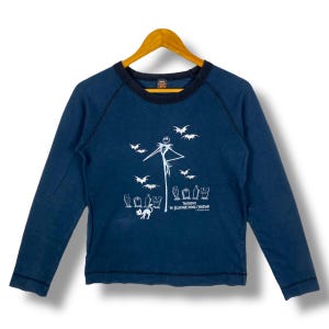 Puede incluir: Camiseta de manga larga azul marino con un gr&aacute;fico blanco de Jack Skellington, murci&eacute;lagos y l&aacute;pidas. El texto "The Nightmare Before Christmas" est&aacute; impreso debajo. La camiseta tiene un cuello azul oscuro y est&aacute; colgada en una percha de madera.