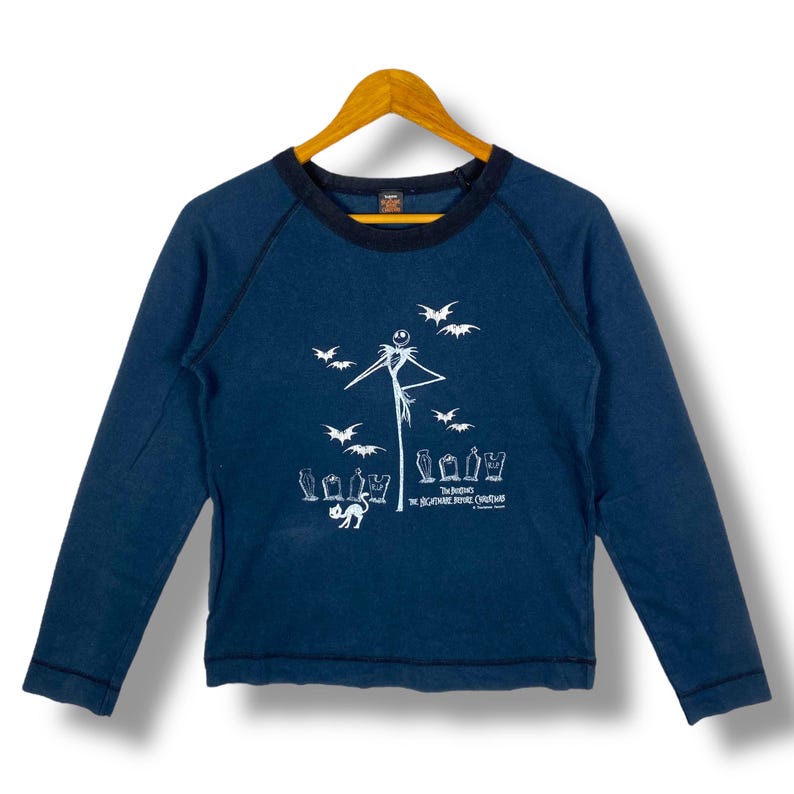 Puede incluir: Camiseta de manga larga azul marino con un gr&aacute;fico blanco de Jack Skellington, murci&eacute;lagos y l&aacute;pidas. El texto "The Nightmare Before Christmas" est&aacute; impreso debajo. La camiseta tiene un cuello azul oscuro y est&aacute; colgada en una percha de madera.