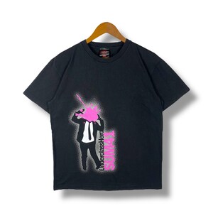 May include: Black t-shirt with a pink and white graphic of a man in a suit with the text "Underclass Hero" and "Shoot" printed on the shirt.