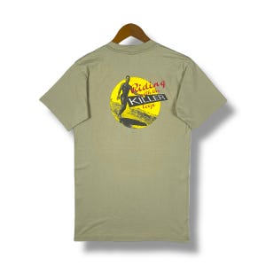 Op de afbeelding: Een lichtgroen T-shirt met korte mouwen met een gele cirkelvormige afbeelding op de rug. De afbeelding toont een surfer die op een golf rijdt en de tekst "Riding with the Killer Loop".