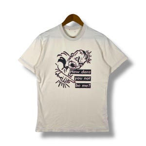 Op de afbeelding: Een wit T-shirt met een zwart-witte illustratie van een persoon met de tekst "How dare you not be me?"