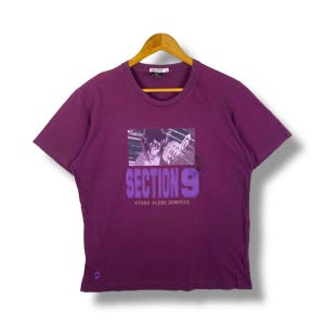 以下が含まれることがあります： アニメ風のイラストと「SECTION 9 STAND ALONE COMPLEX」の文字が入った紫色のTシャツ。木製のハンガーにかかっています。