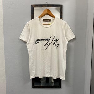 May include: A white short-sleeved t-shirt featuring a black stylized script graphic across the chest. The garment has a crew neckline and a brown interior neck label with the text Ground Y printed in white. The shirt is displayed on a wooden hanger.