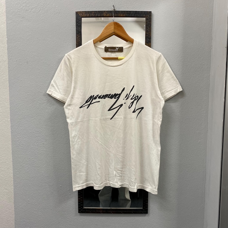 May include: A white short-sleeved t-shirt featuring a black stylized script graphic across the chest. The garment has a crew neckline and a brown interior neck label with the text Ground Y printed in white. The shirt is displayed on a wooden hanger.