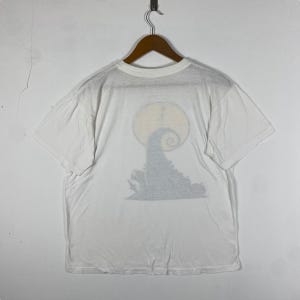 Puede incluir: Camiseta blanca con una impresi&oacute;n descolorida del personaje de Pesadilla Antes de Navidad Jack Skellington frente a una luna llena.