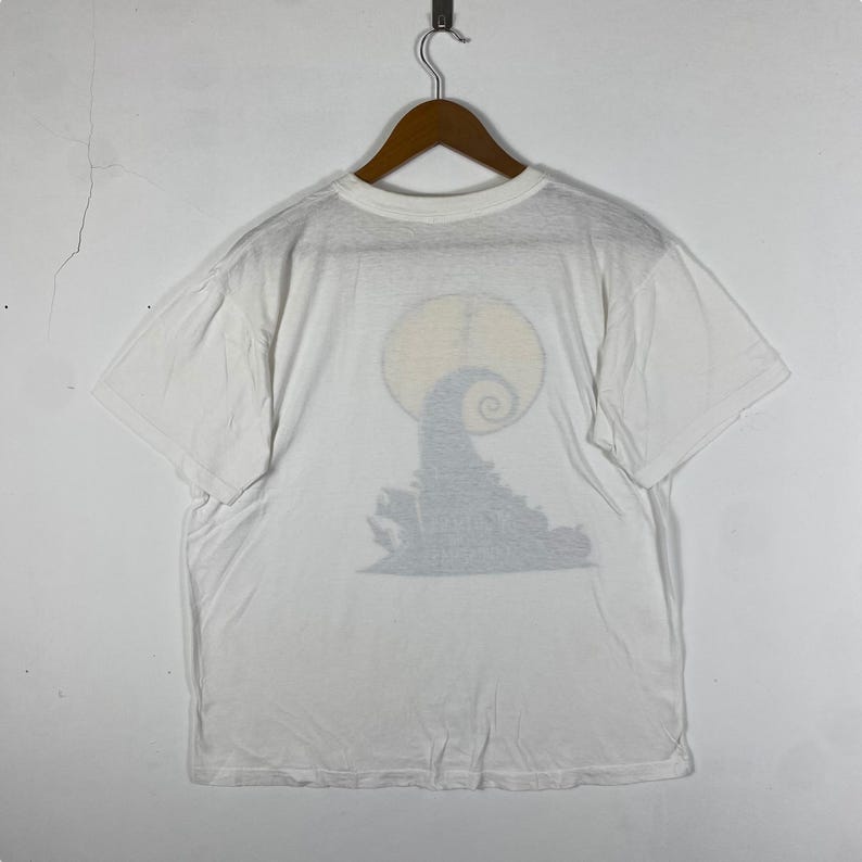 Puede incluir: Camiseta blanca con una impresi&oacute;n descolorida del personaje de Pesadilla Antes de Navidad Jack Skellington frente a una luna llena.