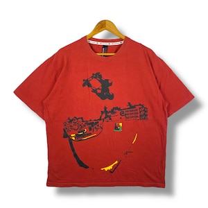 May include: Red t-shirt with a black graphic of a skateboarder performing a trick. The shirt also features abstract shapes and text in black, yellow, and green. The shirt is hanging on a wooden hanger.
