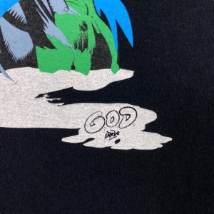 K&ouml;nnte beinhalten: Schwarzes T-Shirt mit einem grafischen Aufdruck einer gr&uuml;nen und blauen Figur mit dem Wort "GOD" in wei&szlig; darunter.