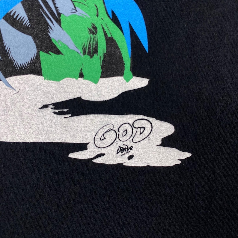 K&ouml;nnte beinhalten: Schwarzes T-Shirt mit einem grafischen Aufdruck einer gr&uuml;nen und blauen Figur mit dem Wort "GOD" in wei&szlig; darunter.