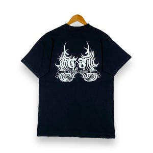 以下が含まれることがあります： 黒の半袖Tシャツ。背面に白い部族模様と「Christian Fletcher」の文字が入っています。