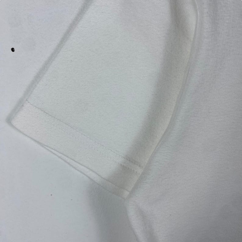 May include: Close-up of the white fabric of a t-shirt, showing the stitching along the edge of the sleeve.