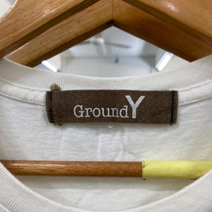 May include: A white t-shirt featuring a rectangular brown fabric neck label with the text Ground Y embroidered in white thread. The label is stitched onto the inner collar of the garment, which is positioned on a wooden hanger.