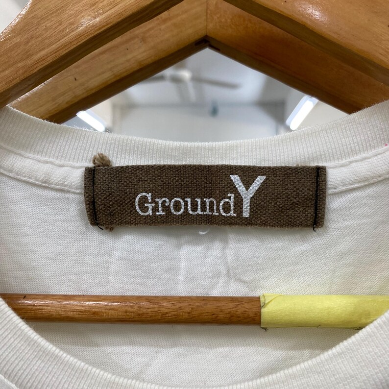 May include: A white t-shirt featuring a rectangular brown fabric neck label with the text Ground Y embroidered in white thread. The label is stitched onto the inner collar of the garment, which is positioned on a wooden hanger.