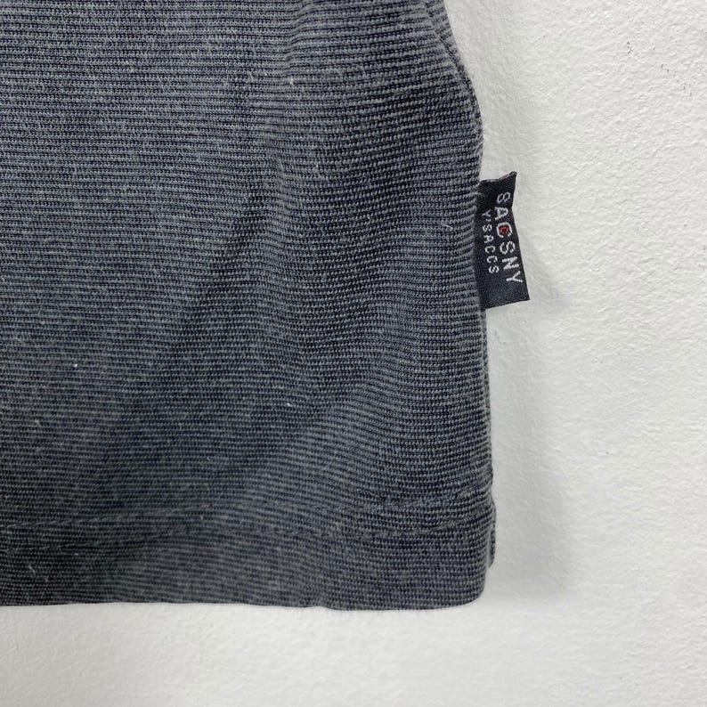 K&ouml;nnte beinhalten: Ein dunkelgraues geripptes T-Shirt mit einem schwarzen Etikett, auf dem rechts unten "SACSNY" steht.