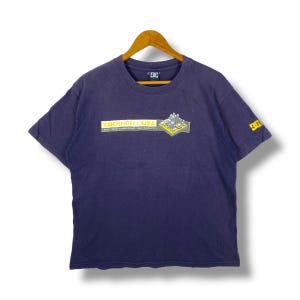 May include: A navy blue short-sleeved t-shirt with a crew neck. The shirt features a graphic on the front with the text "DCSHOECOUSA" and a geometric design in yellow and gray. A smaller logo is on the sleeve.