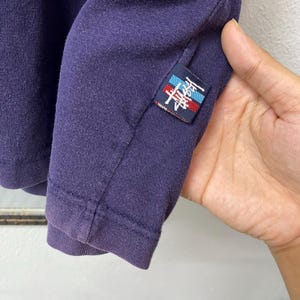 May include: Close-up of a dark purple long-sleeved shirt. A small rectangular patch with a logo in red, white, and blue is sewn on the sleeve. The shirt's fabric appears to be a soft knit material.