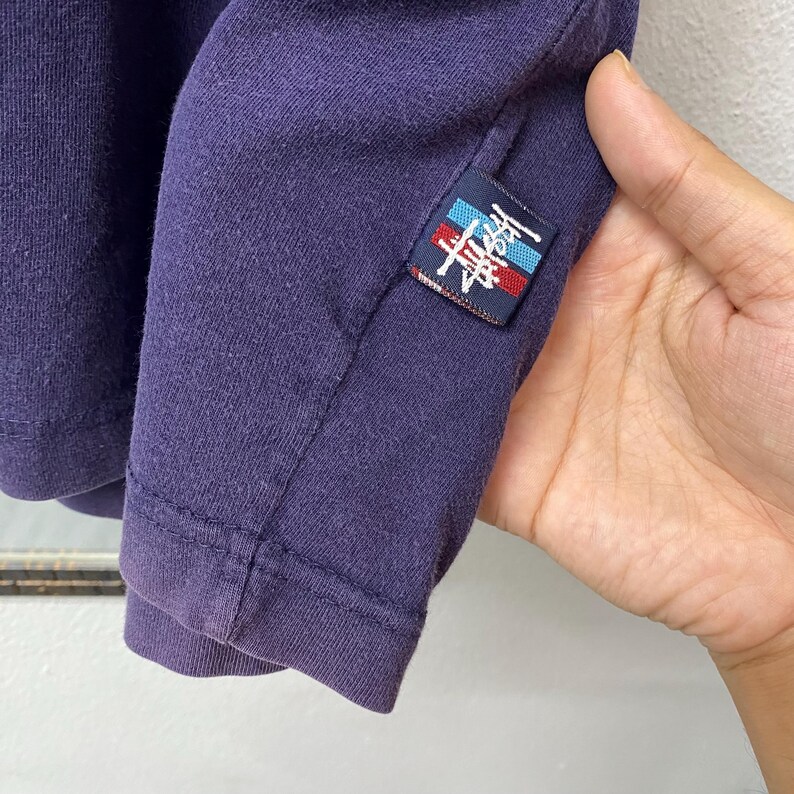 May include: Close-up of a dark purple long-sleeved shirt. A small rectangular patch with a logo in red, white, and blue is sewn on the sleeve. The shirt's fabric appears to be a soft knit material.