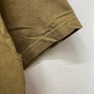 May include: Close-up of a section of a mustard-colored t-shirt. The sleeve's hem is visible, with a detailed seam. The fabric appears to be a soft, textured material. The background is a plain, off-white wall.