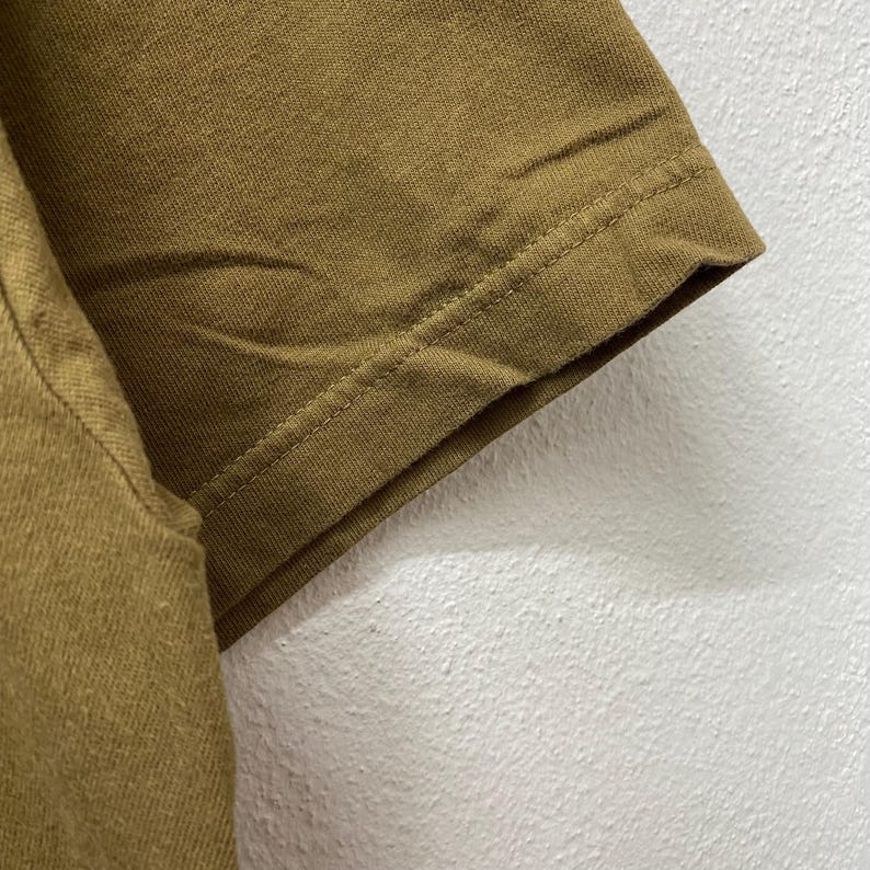 May include: Close-up of a section of a mustard-colored t-shirt. The sleeve's hem is visible, with a detailed seam. The fabric appears to be a soft, textured material. The background is a plain, off-white wall.