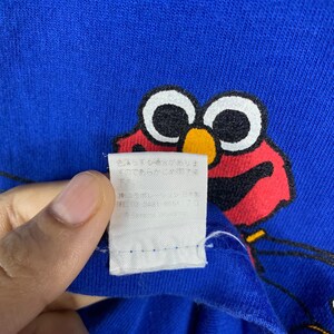 May include: A blue t-shirt featuring a red Elmo character graphic. The tag on the shirt reads "Sesame Street".