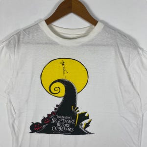 Puede incluir: Camiseta blanca con un gr&aacute;fico negro y amarillo de Jack Skellington de la pel&iacute;cula de Tim Burton Pesadilla antes de Navidad. El gr&aacute;fico muestra a Jack de pie en una colina con una luna amarilla detr&aacute;s de &eacute;l. El texto "Tim Burton's Nightmare Before Christmas" est&aacute; impreso debajo del gr&aacute;fico.