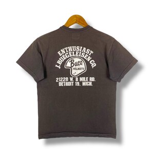 May include: Dark gray t-shirt with a white graphic on the back. The graphic includes the text "ENTHUSIAST BUEGELEISEN CO." and "Buco HELMETS" with an image of a helmet. Below the graphic is the text "21220 W. 8 MILE RD. DETROIT 19. MICH."