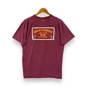 May include: A maroon short-sleeved t-shirt with a red rectangular patch on the back that says "Abercrombie and Fitch Est. 1892 Surfboards" in yellow lettering.